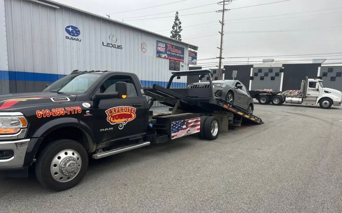 Black pickup truck towing a damaged silver car on a flatbed trailer outside an auto shop.