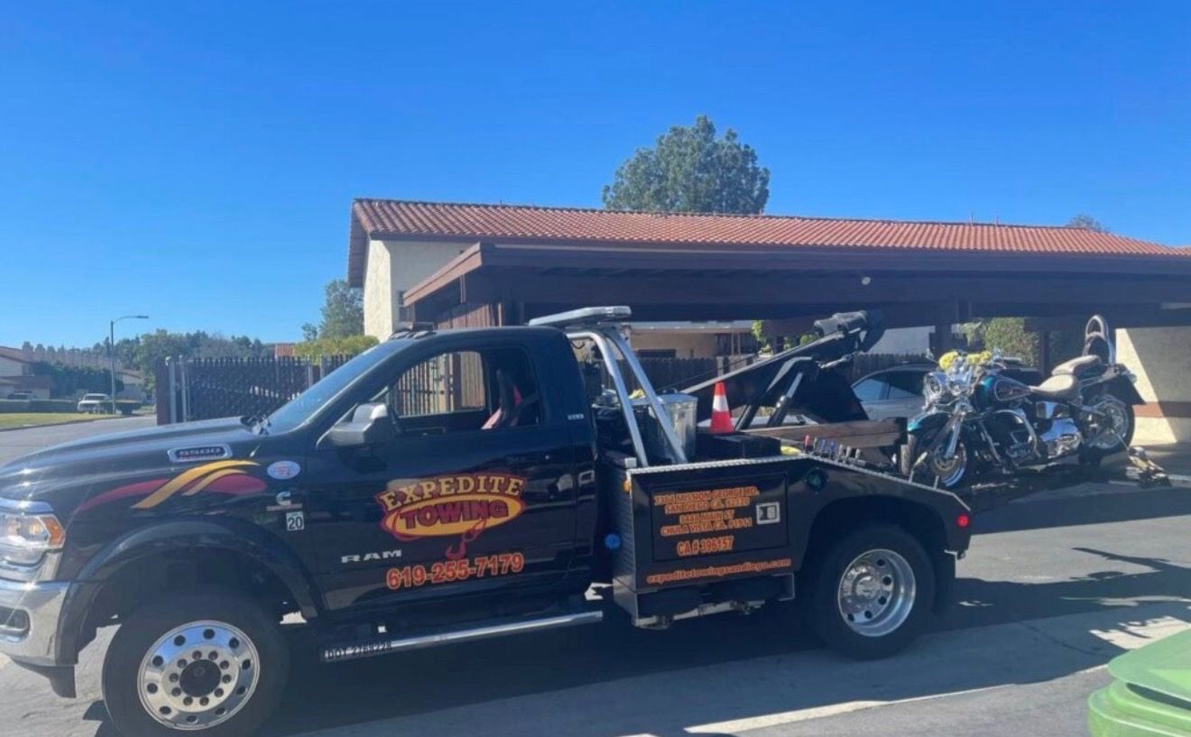Black tow truck carrying motorcycles in a sunny driveway or parking area.