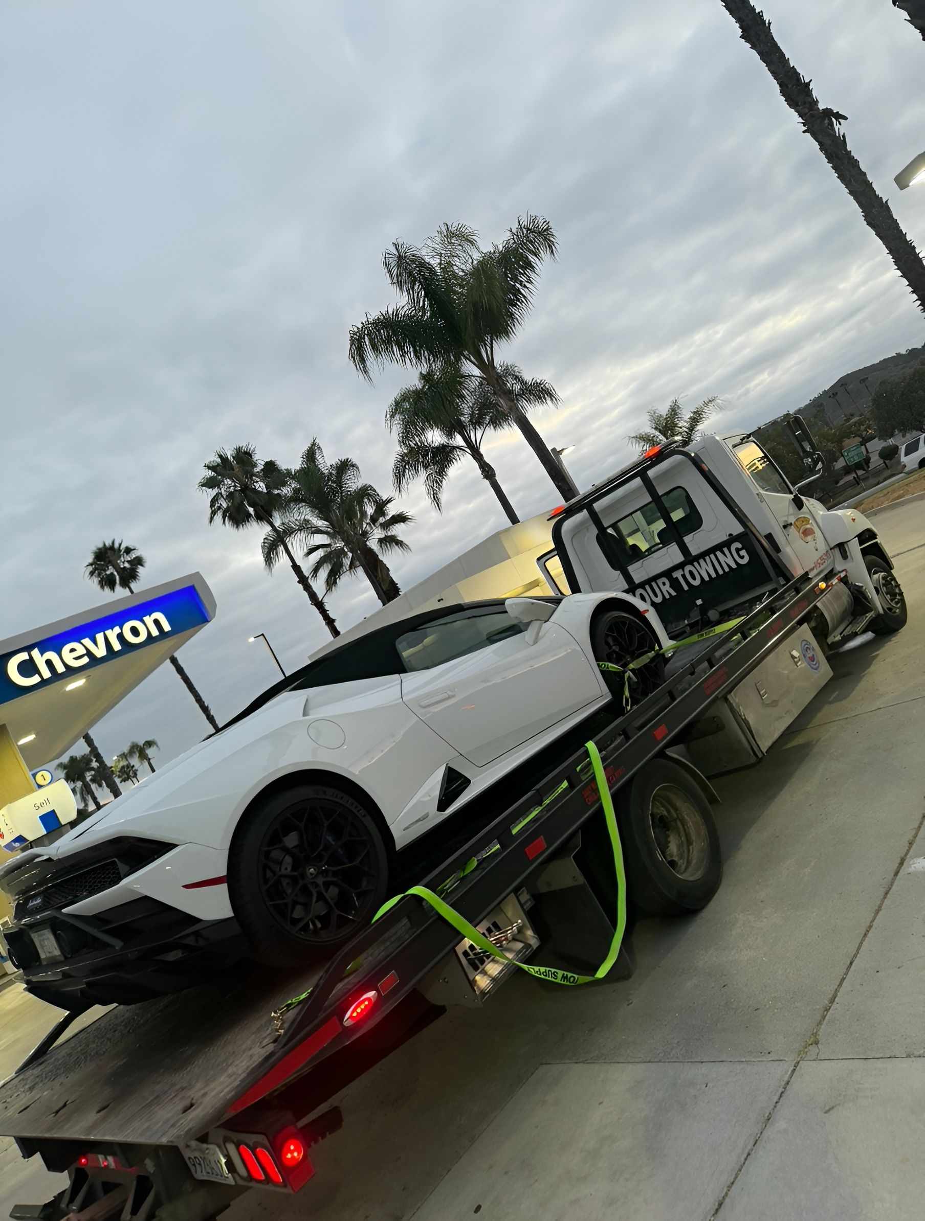 White sports car on a tow truck at a Chevron gas station under cloudy skies.