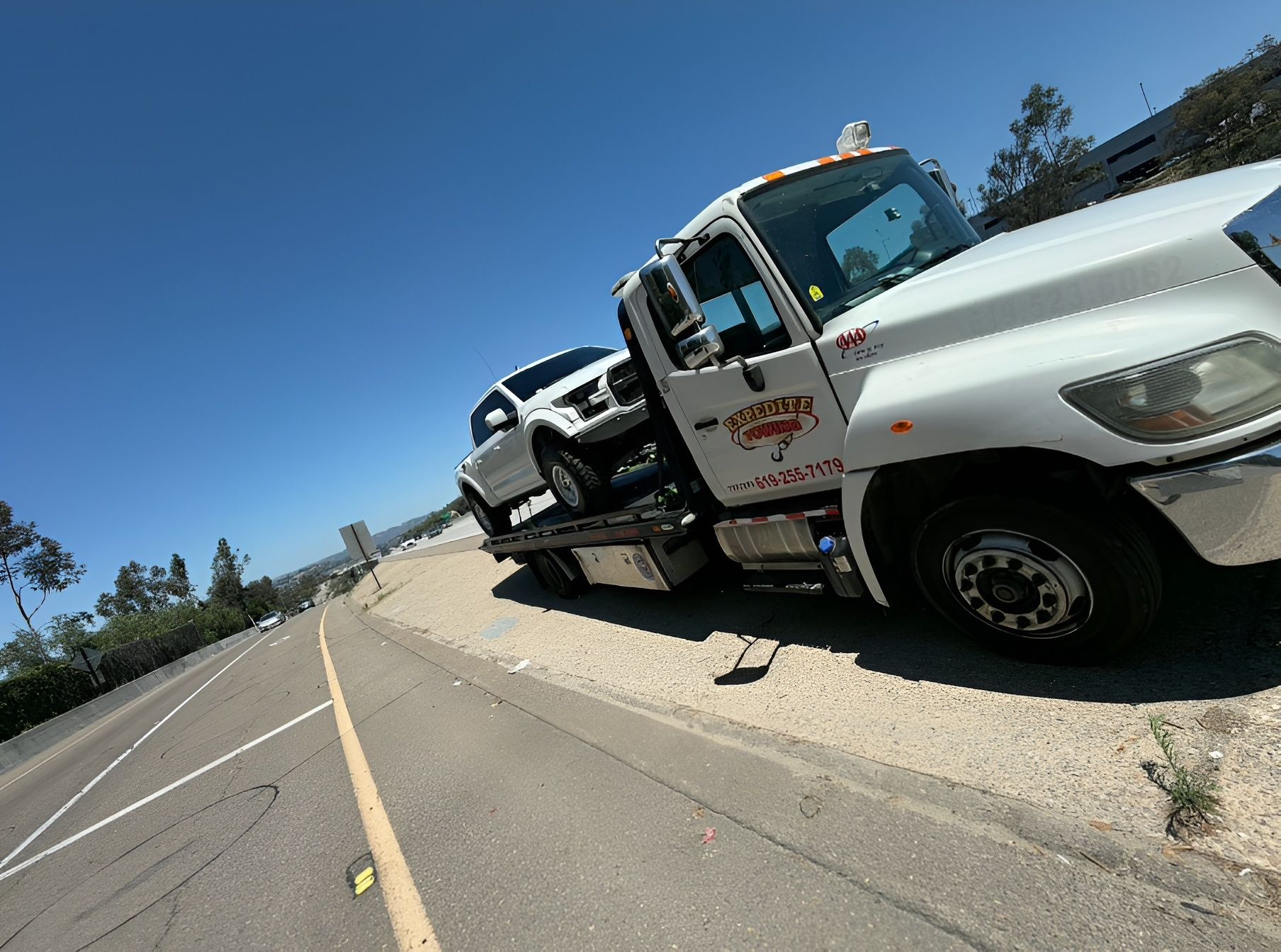 Tow truck hauling a white car on a roadside beside an empty highway under a clear blue sky.