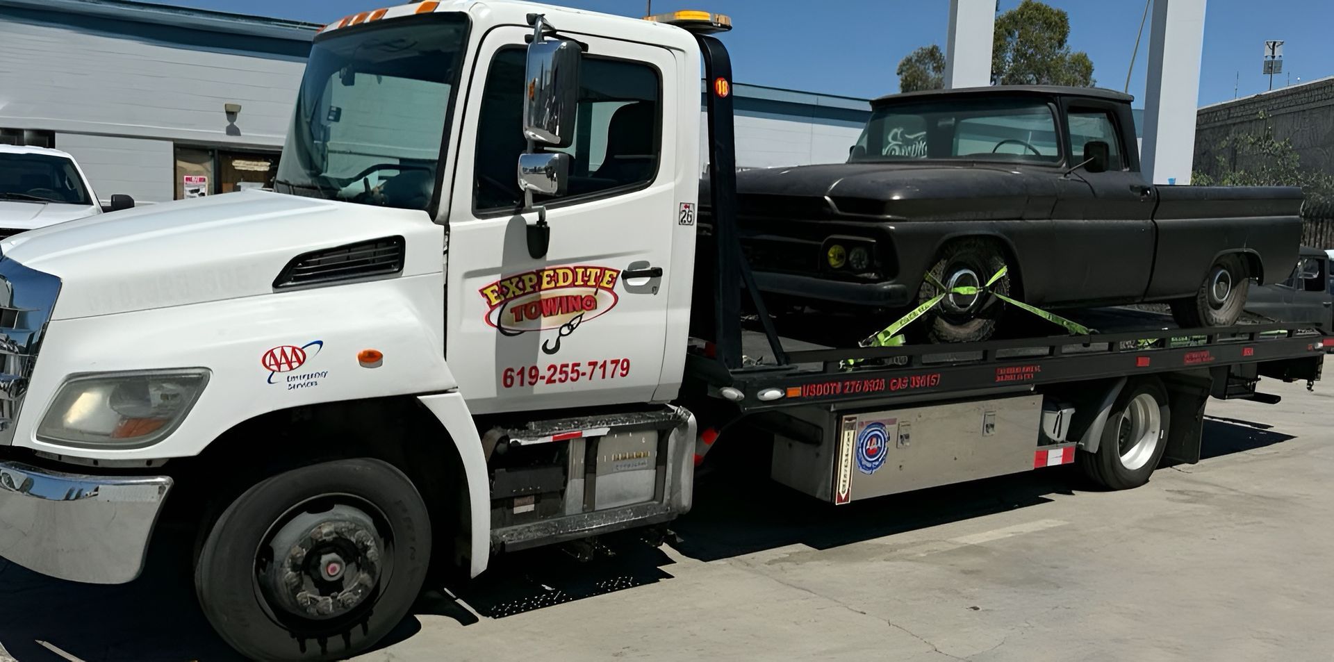 White tow truck hauling a black pickup on a flatbed in a sunny parking lot.