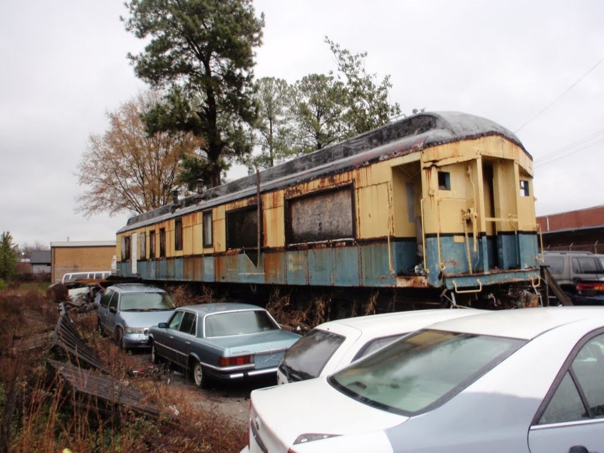 Old, rusted train car parked outdoors beside several cars in a scrapyard.