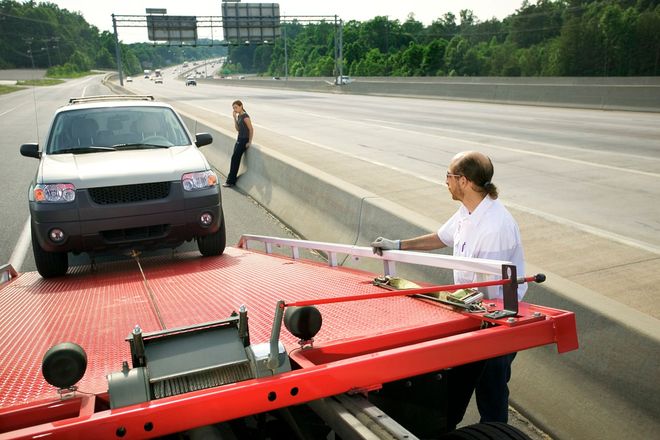 Tow truck towing a black SUV on a highway shoulder, with a man standing beside the car and driver at the truck controls.
