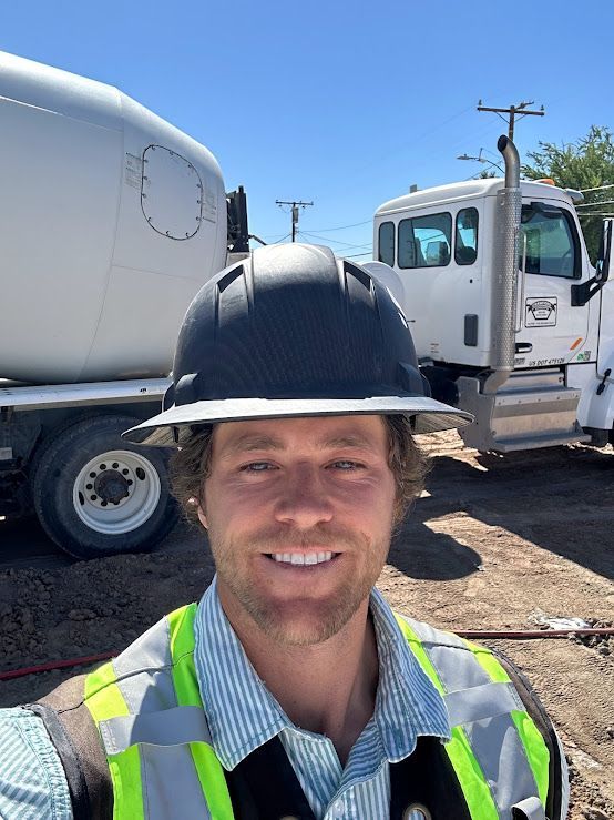 A construction worker in a hard hat and reflective vest smiles for a selfie with a concrete truck in the background.