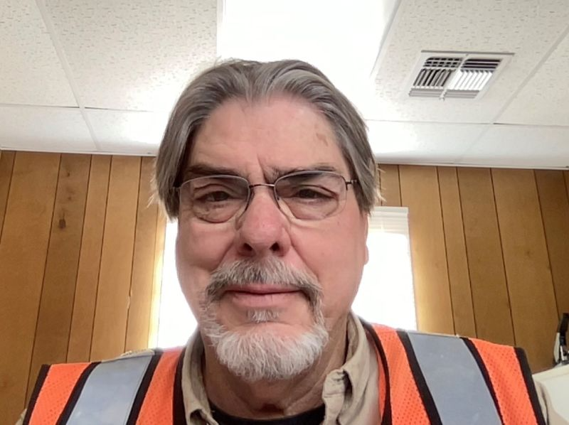 Man wearing glasses and an orange safety vest, standing indoors in front of wood paneling, looking at the camera.