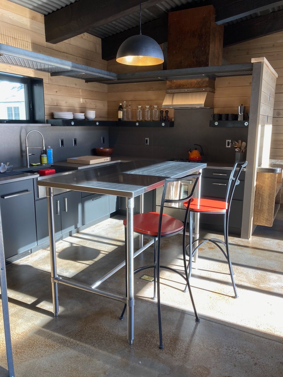 Outdoor kitchen with dark gray cabinets, stainless steel island, red chairs, and corrugated metal roof.