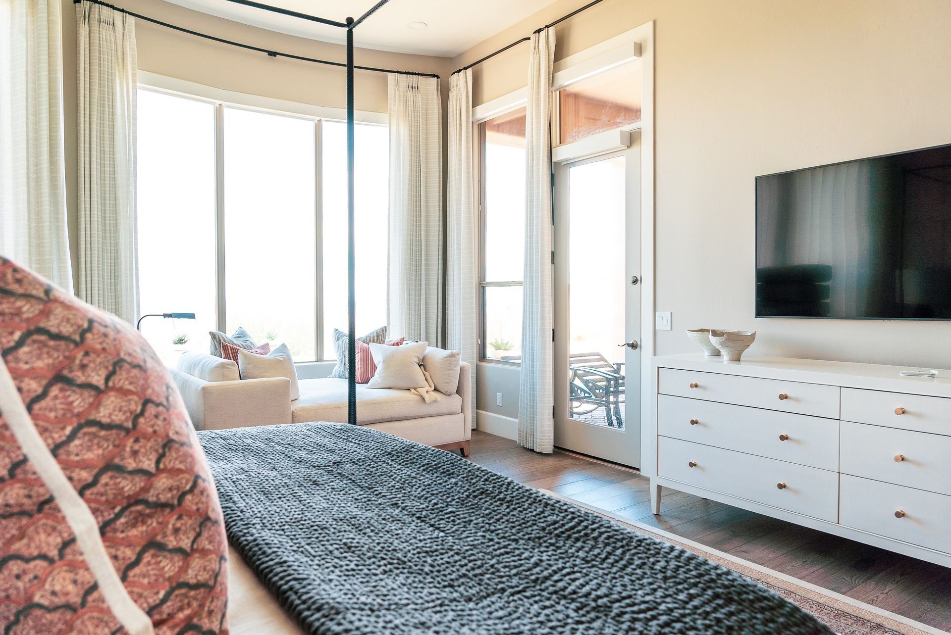 A bedroom with a canopy bed , dresser , television and large windows.