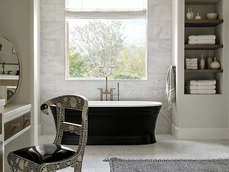 A bathroom with a black tub , a chair , a mirror and a window.
