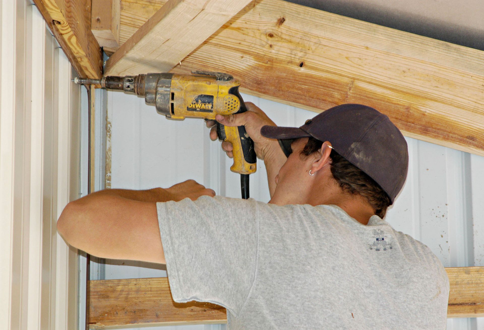 Person using a yellow drill to attach wooden beams inside a white-walled structure.