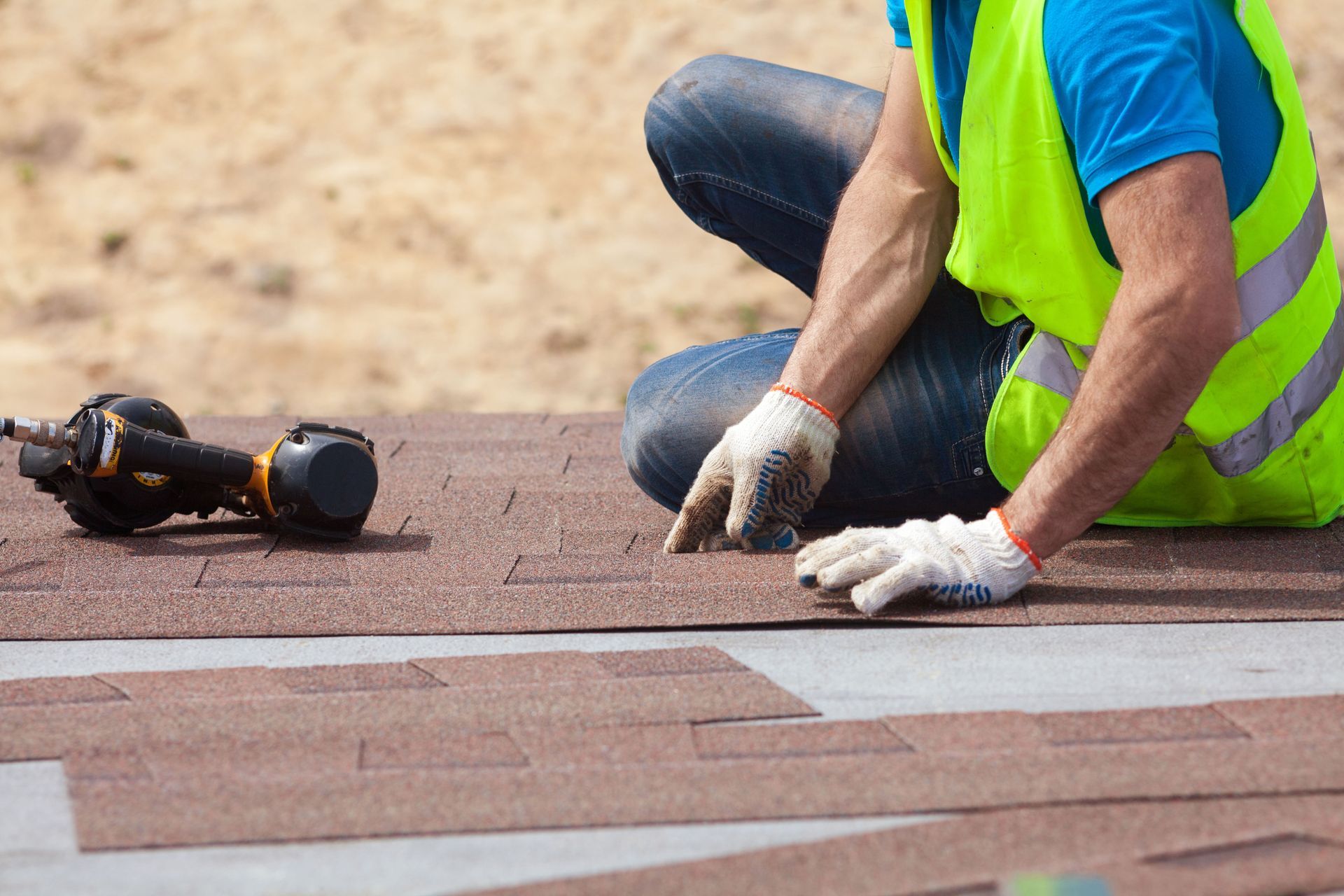 Roofer in safety vest installing brown shingles on a roof with a drill and gloves.