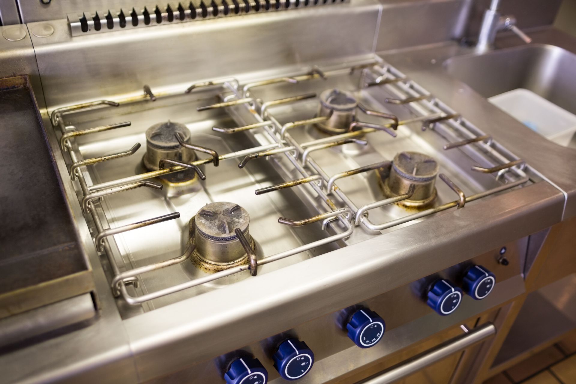 Stainless steel commercial gas stovetop with four burners, blue knobs.