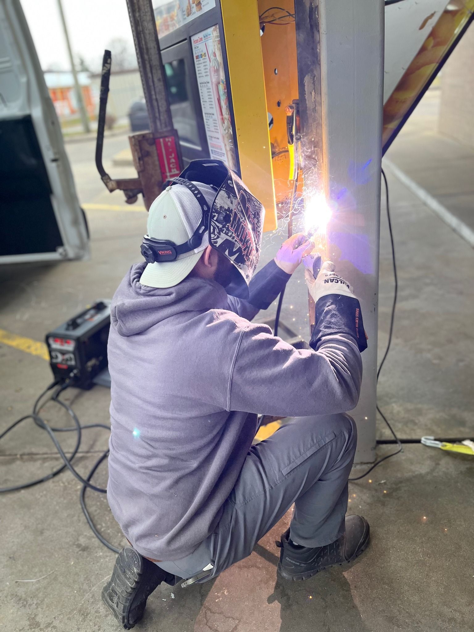 Welder kneeling, welding metal structure outdoors. Sparking. Gray sweatshirt, camo helmet. Welding machine nearby.