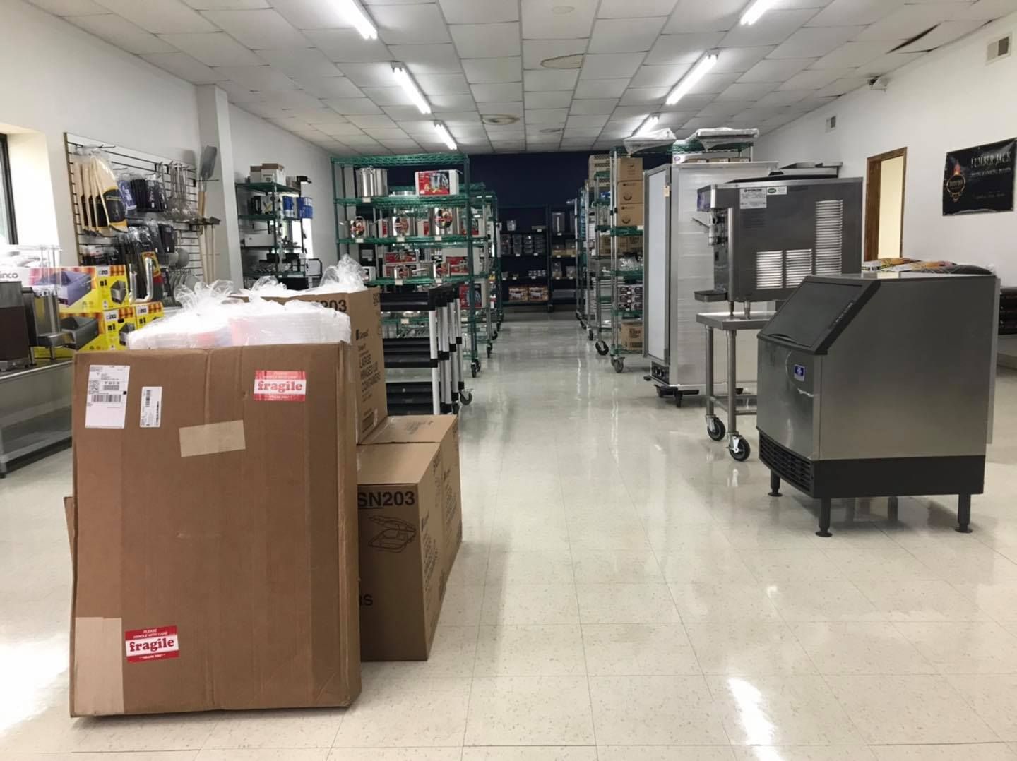 Inside a supply store, shelves of kitchen equipment line the aisles. Cardboard boxes are stacked in the foreground.