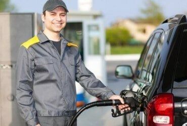 Gas station attendant in gray uniform fueling a black SUV, smiling at the pump.