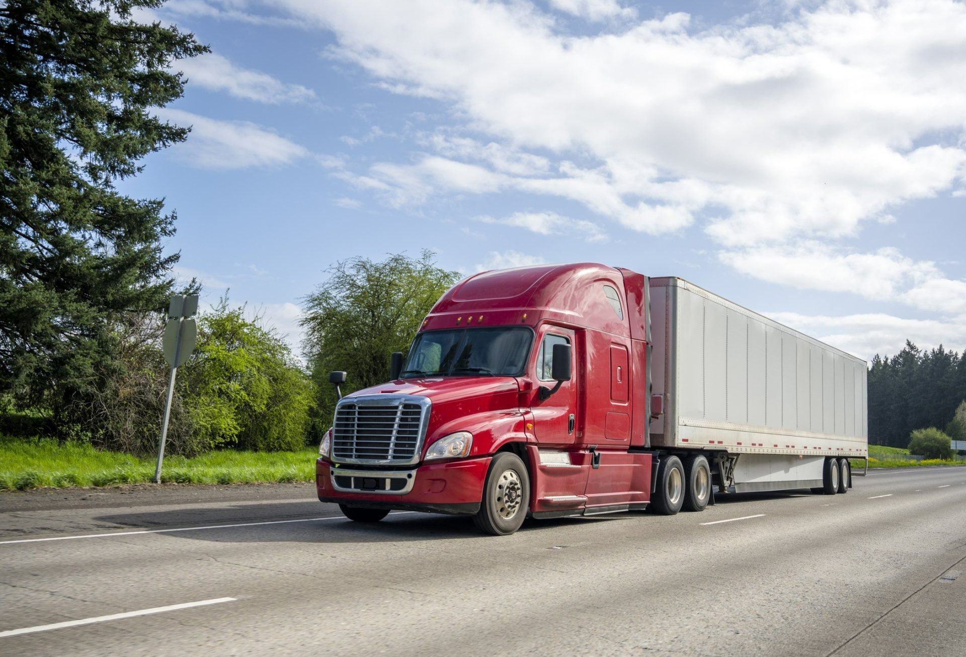Red semi-truck hauling a white trailer on a highway beside trees under a partly cloudy sky