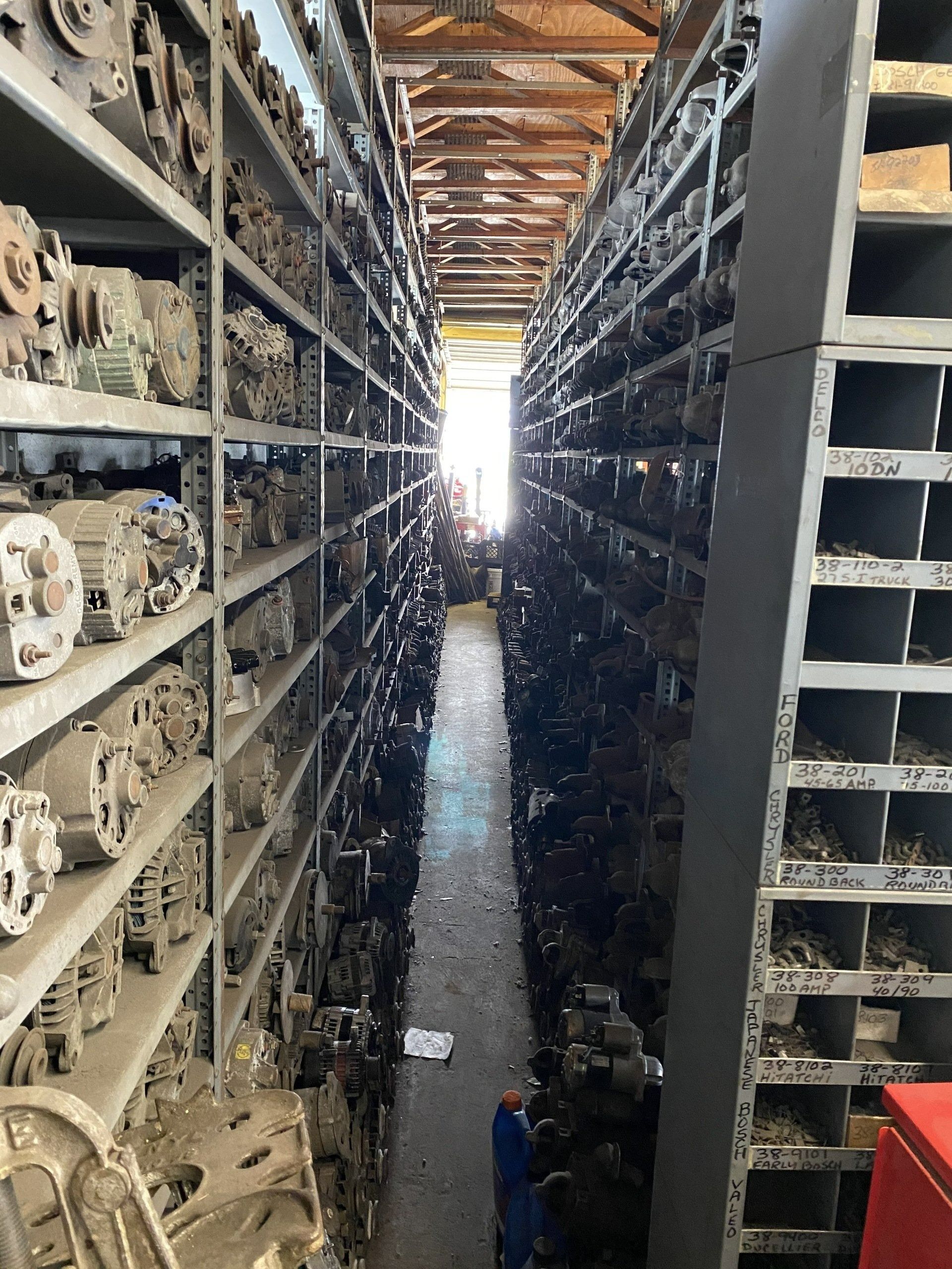 A narrow aisle in a warehouse lined with shelves of old car parts and metal components.
