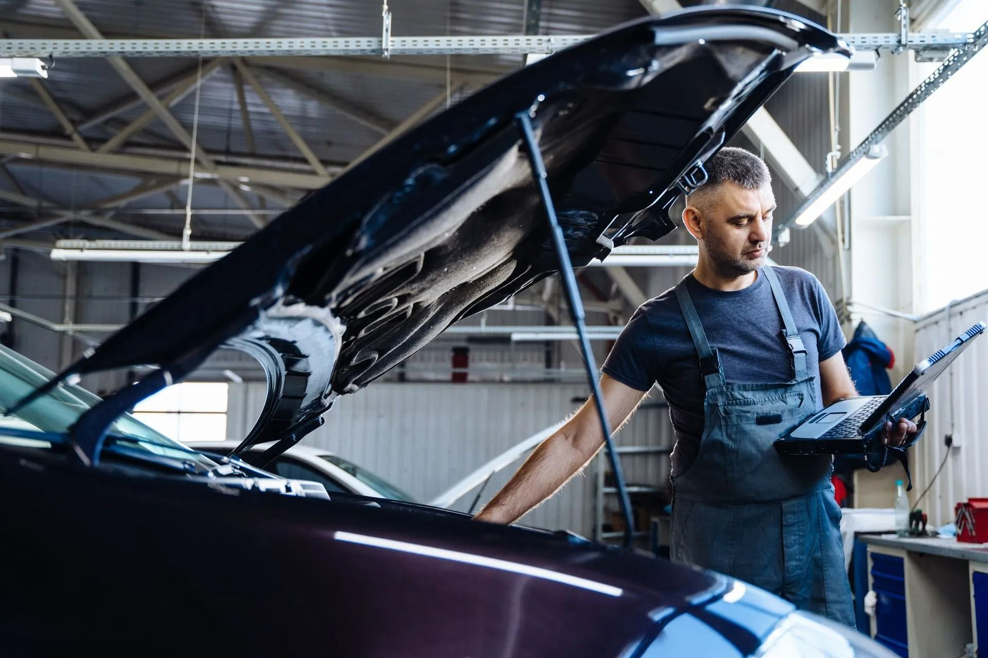 Mechanic inspecting a car engine in an auto repair shop, hood open and diagnostic tablet in hand
