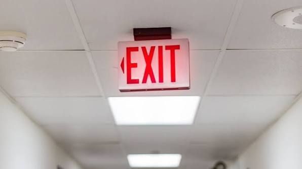 Red EXIT sign hanging from a ceiling in a hallway with fluorescent lights.
