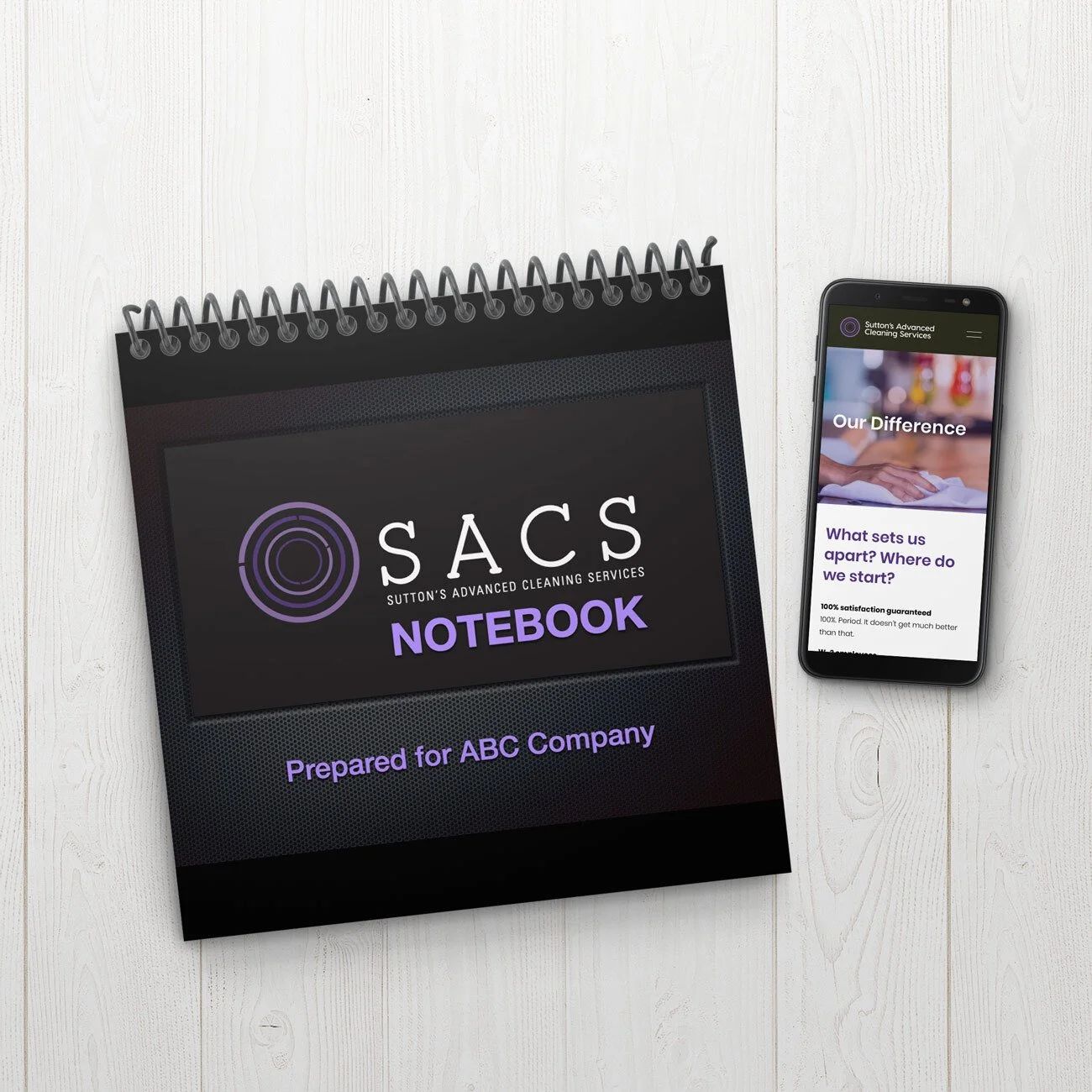 Black spiral notebook labeled “SACS NOTEBOOK” beside a smartphone on a white surface.