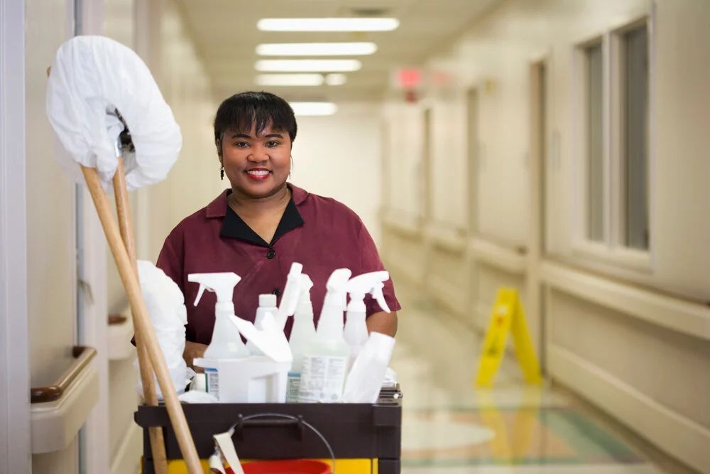 Custodian pushing cleaning cart in a bright hallway, holding a mop with a white cloth head