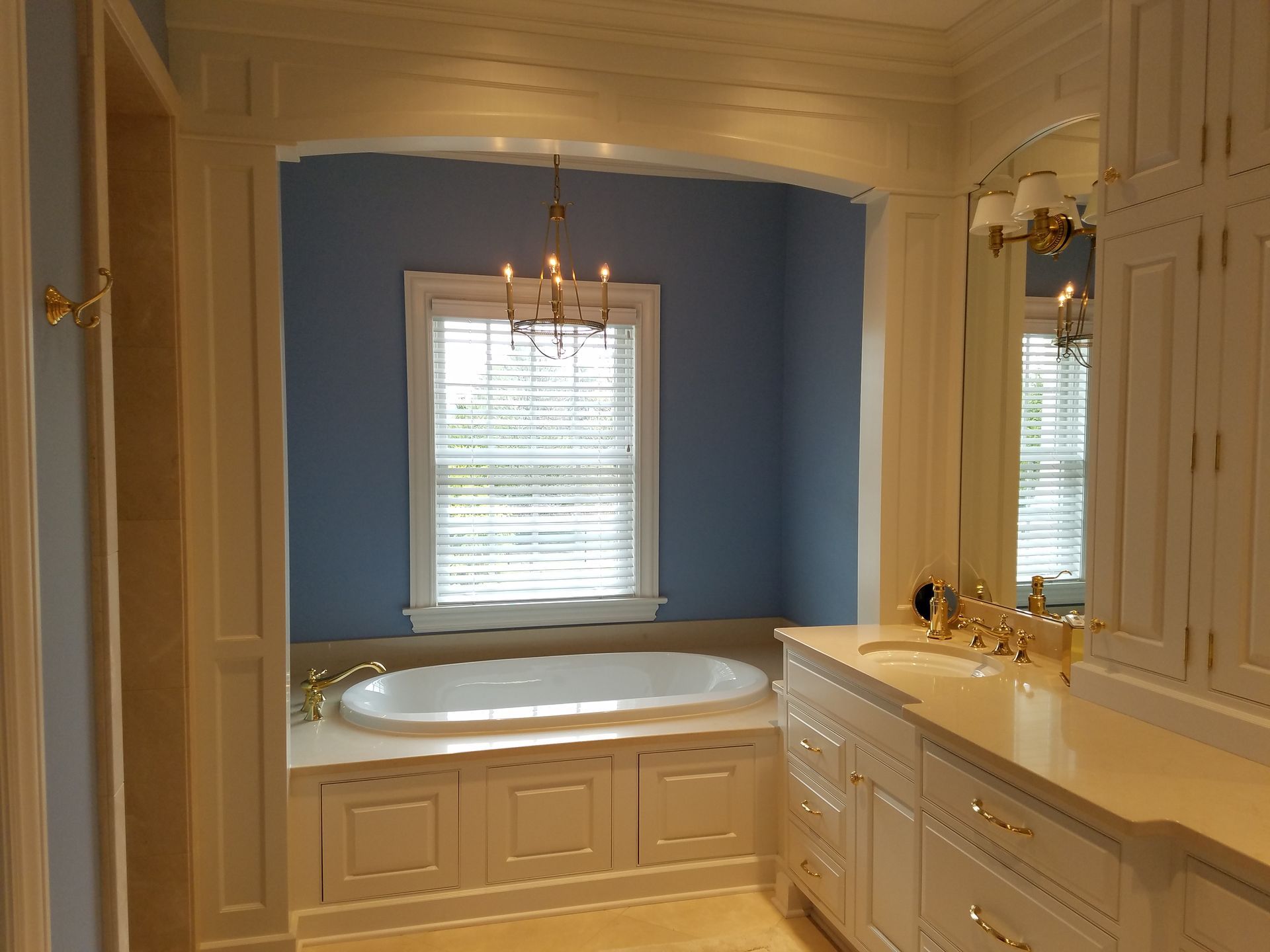 Bathroom with a bathtub, vanity, and window with blinds. Blue walls, white cabinets, and gold fixtures.