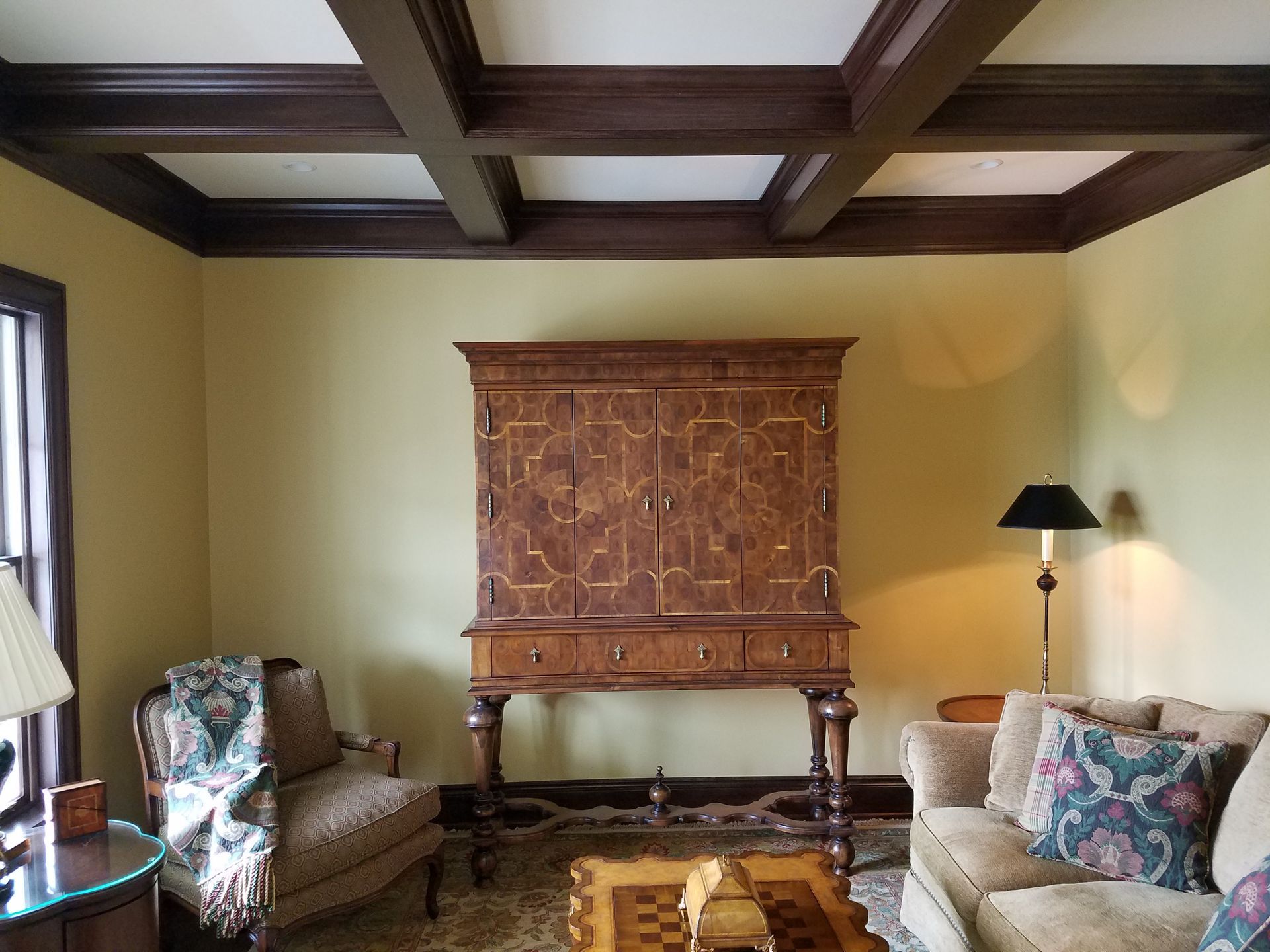 Room with ornate wooden cabinet, sofa, chair, and coffered ceiling.