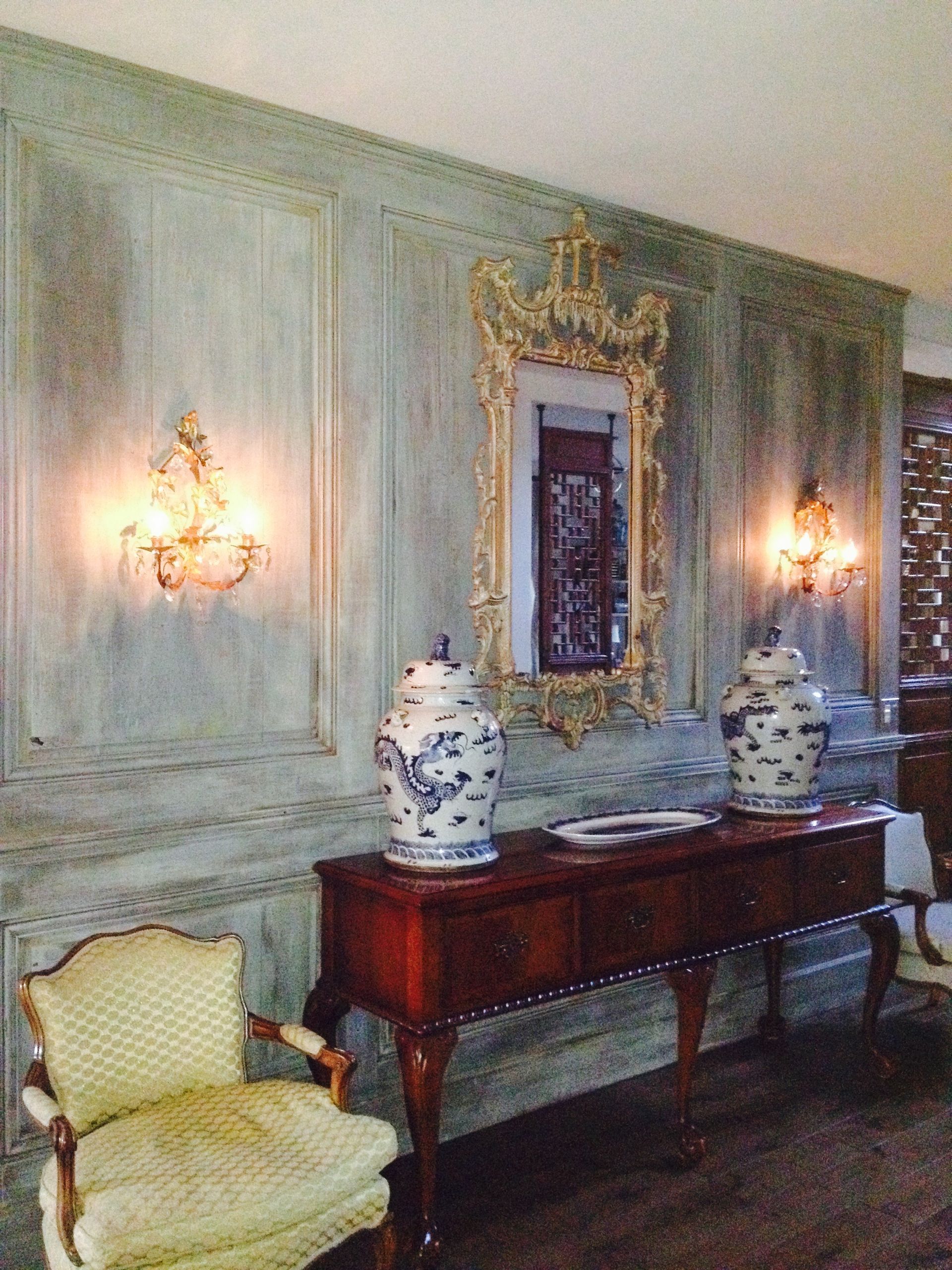Elegant room with antique furniture, gold mirror, blue-and-white vases, and sconces on a textured green wall.