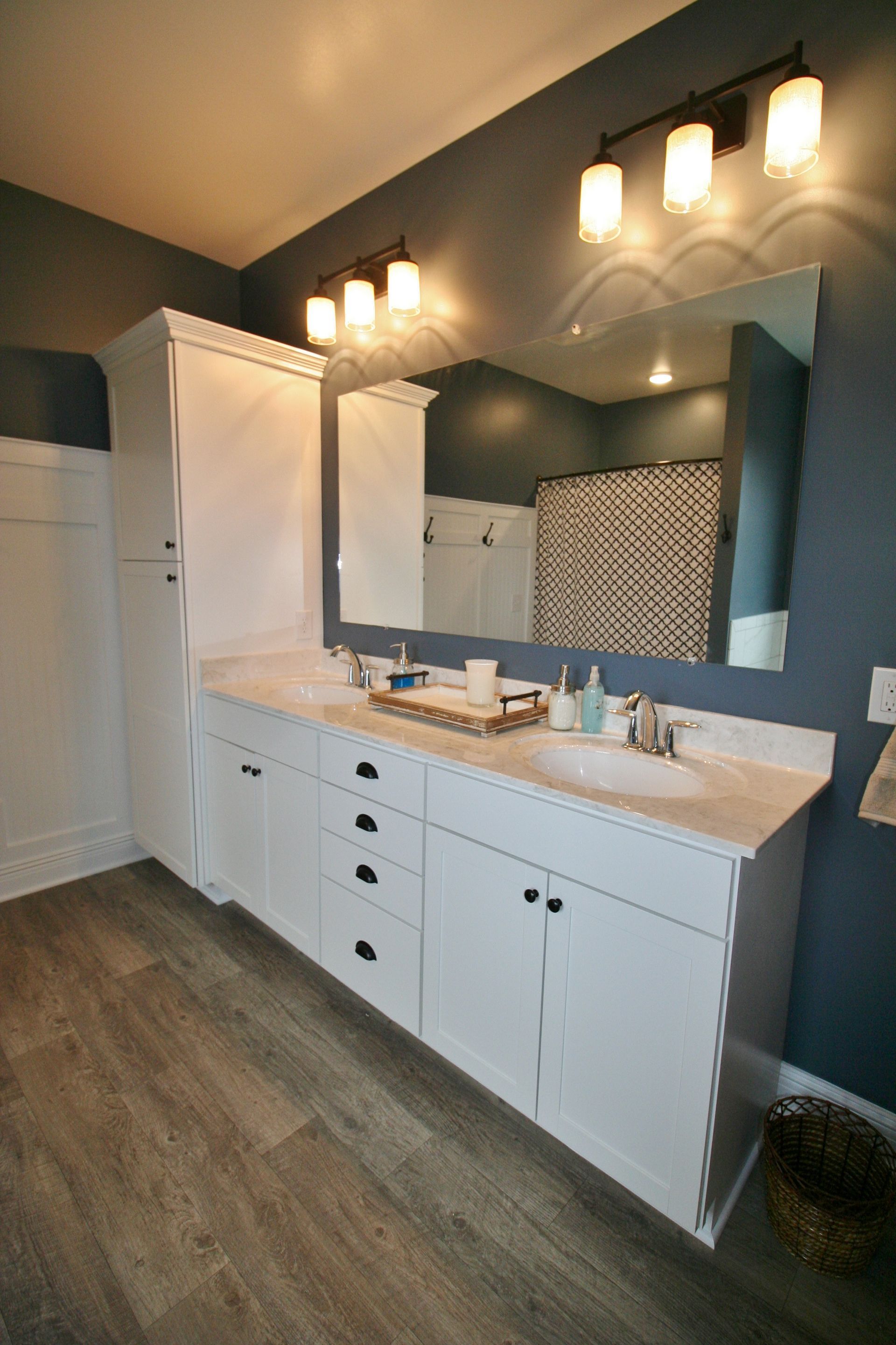 White bathroom vanity with storage tower, double sinks, large mirror, black hardware, and gray walls.
