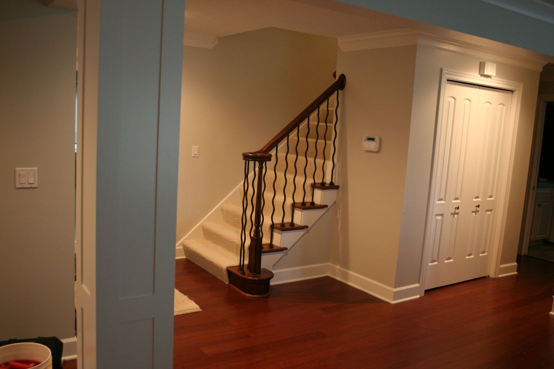 Staircase with dark wood railing and white steps, next to a white door in a tan-colored hallway, with hardwood floors.