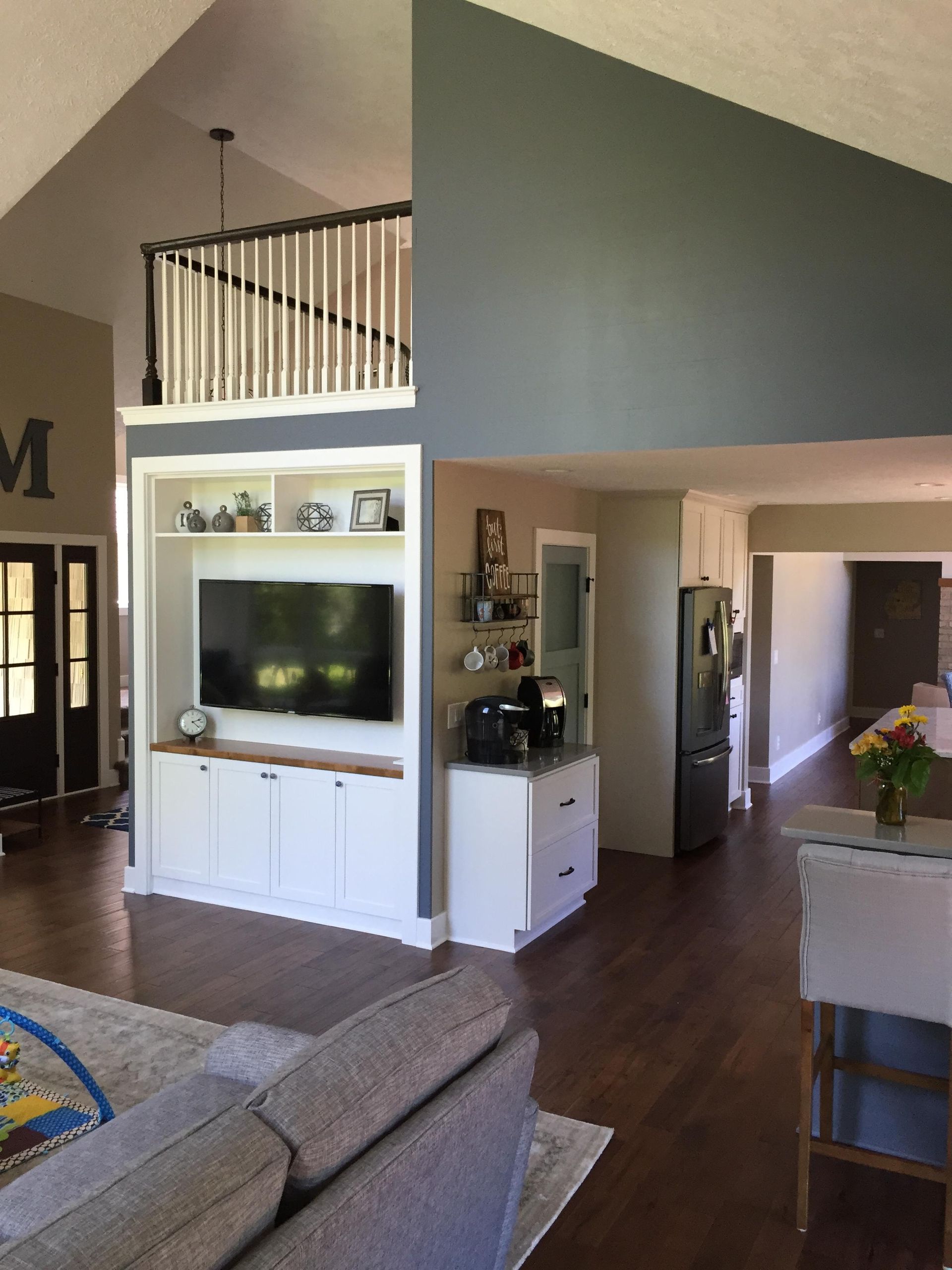 Living room with built-in entertainment center, kitchen in background. White cabinets, TV, coffee bar, and dark wood floors.