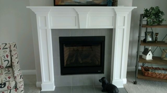 White fireplace with black insert, gray tile, art above. Side bookshelf and chair visible.
