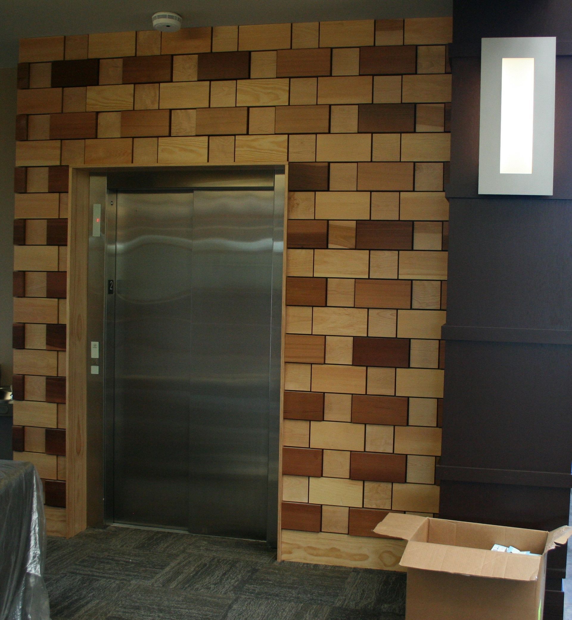 Elevator with stainless steel door framed by a textured wood wall. White wall sconce to the right, cardboard box on the floor.