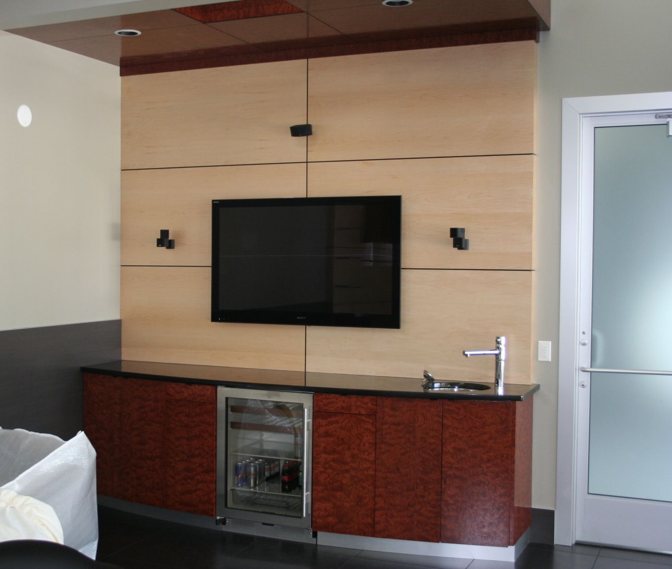 Built-in bar with TV, mini-fridge, and sink. Wood paneling in a modern room with a frosted glass door.