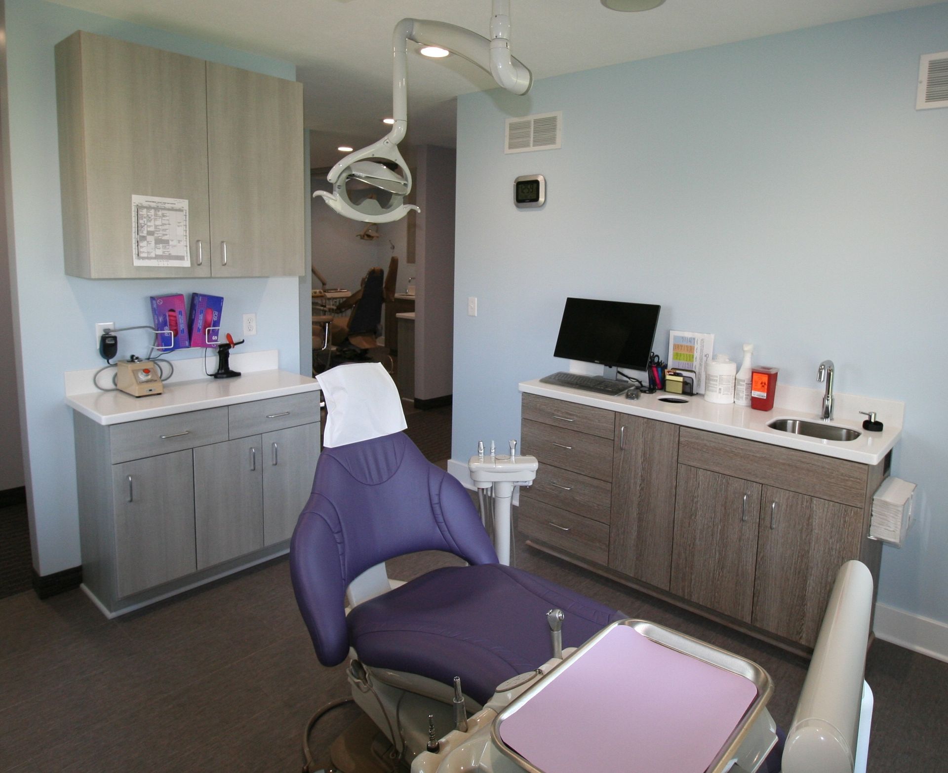 Dental examination room, gray cabinets, light blue walls, purple chair, equipment.