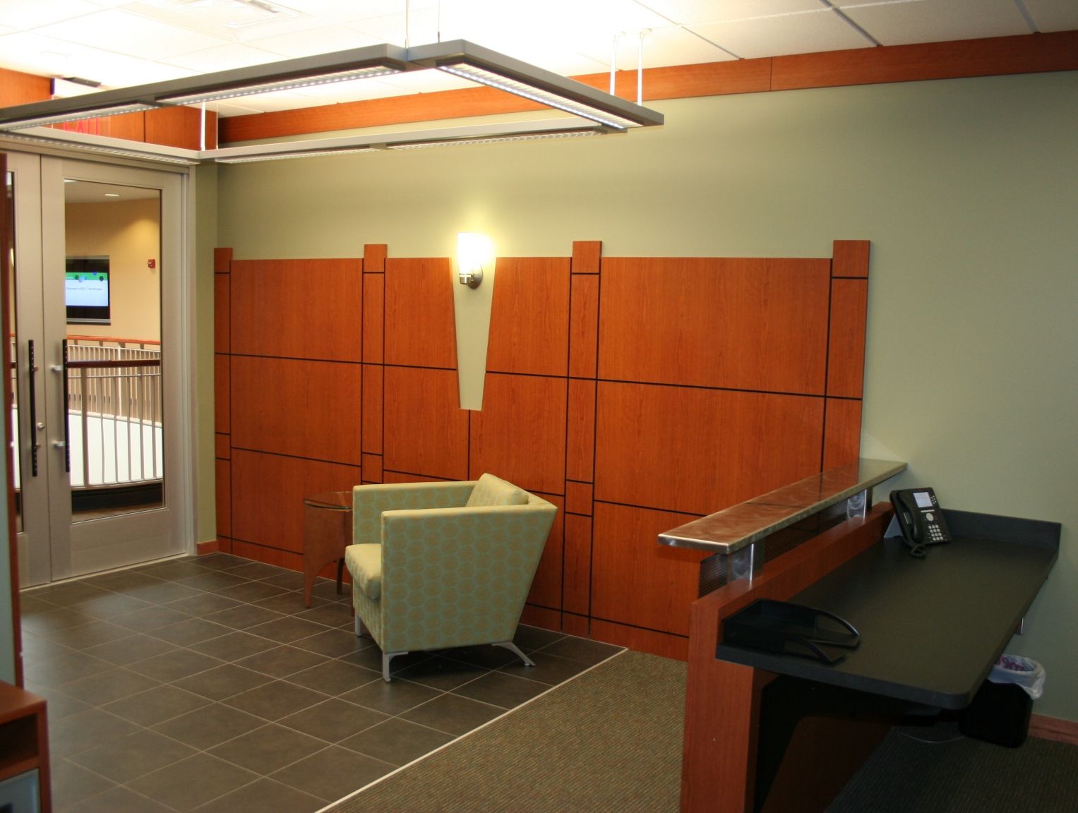 Lobby with wooden wall, armchair, and reception desk. Glass door on left. Brown and green colors.
