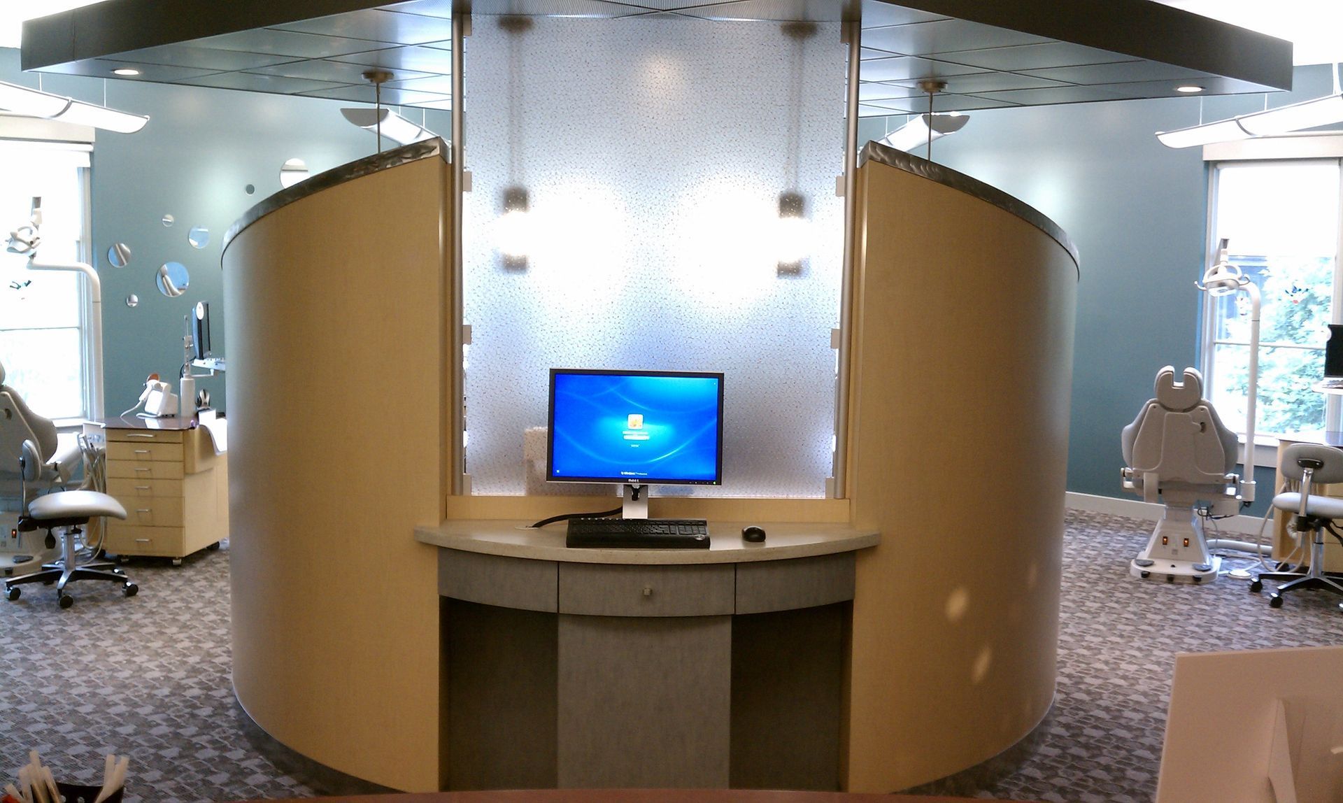 Dental office with computer desk inside a curved booth, light blue walls, carpet, and medical equipment.