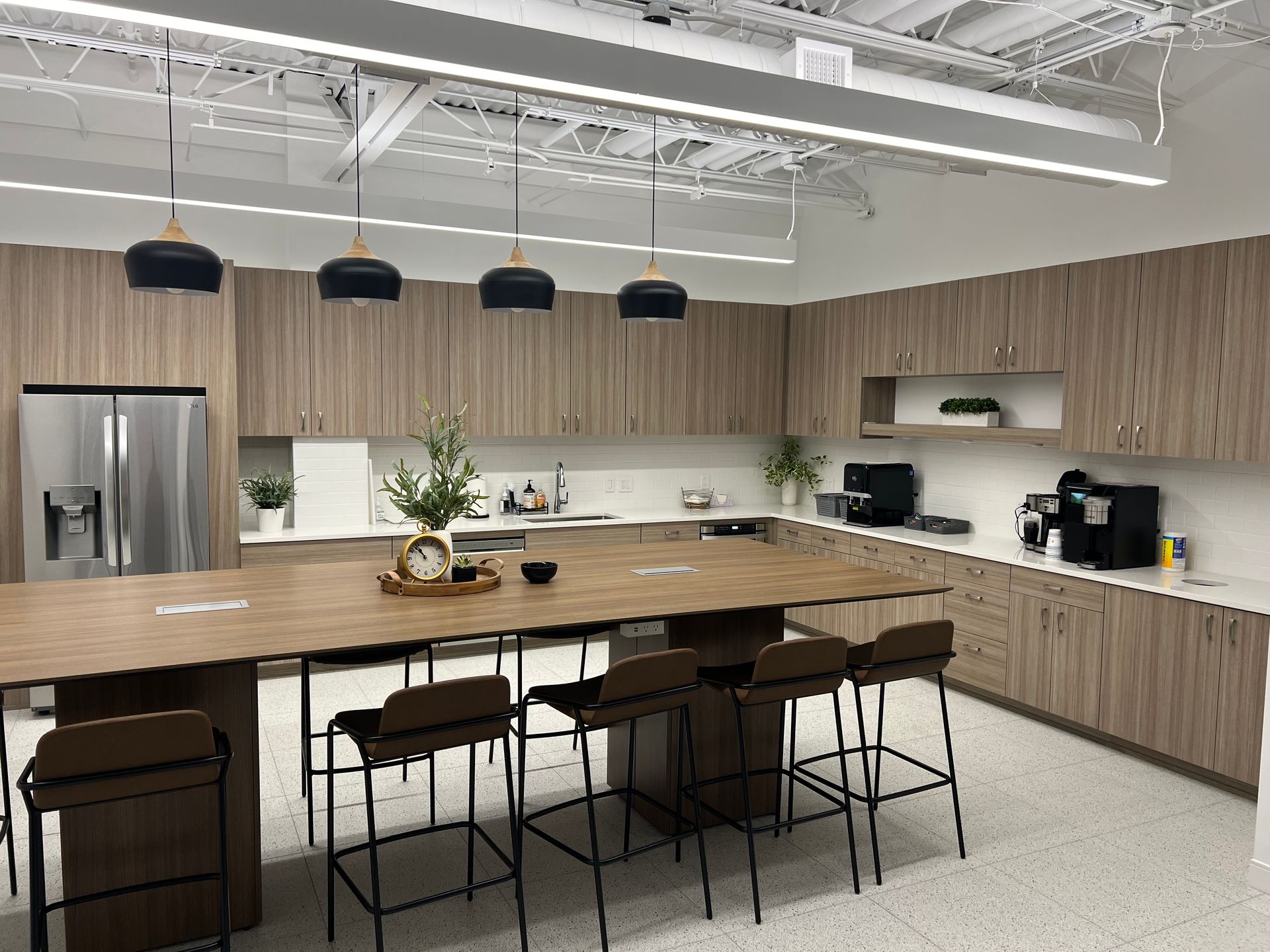 Modern office kitchen with wood cabinets, island seating, pendant lights, and stainless steel appliances.