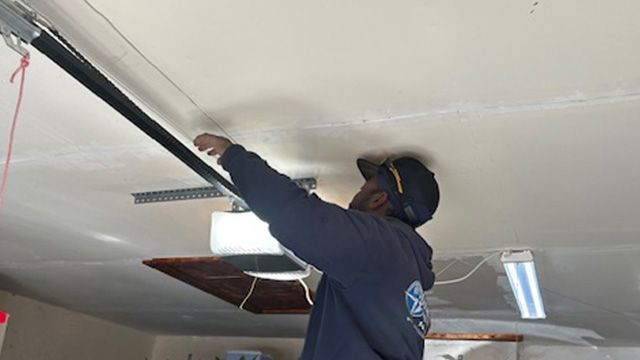 Person working on a garage door spring, indoors. They are wearing a hat and long-sleeved shirt.