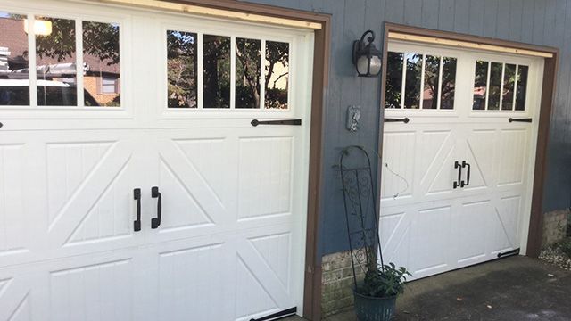 Two white garage doors with windows and black handles, next to a blue wall.