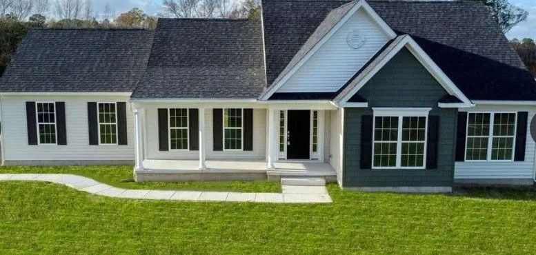 A white house with black shutters, green siding, and a walkway on a grassy lawn.