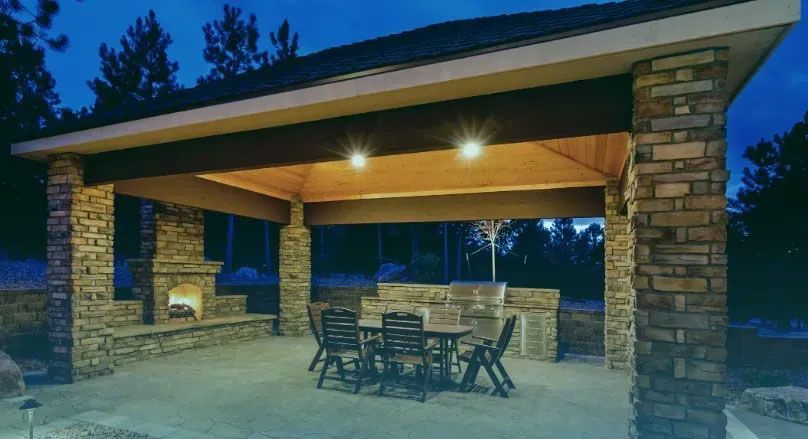 Outdoor kitchen and dining area under a stone-columned pavilion lit at night. Features fireplace, grill, table, and chairs.