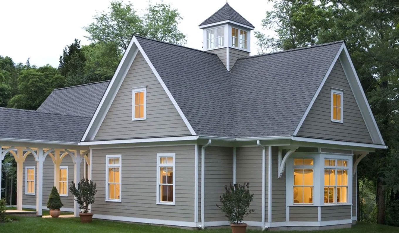 Gray house with a dark gray roof, white trim, and a cupola. Lush green trees in the background.