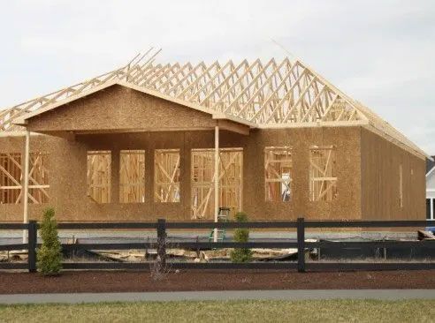 House under construction; wooden frame and roof trusses visible, cloudy sky in background.
