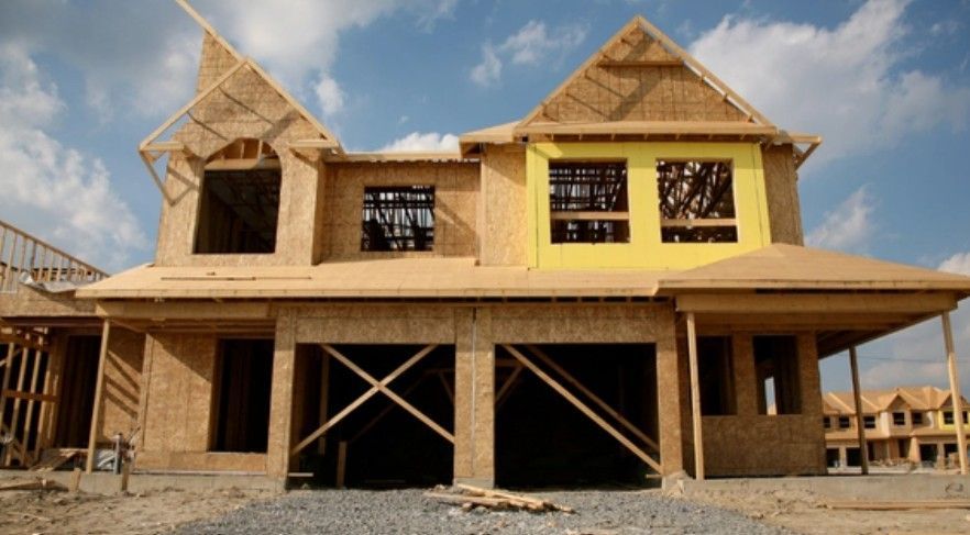 Two-story house under construction with exposed wooden framing and yellow insulation around window openings.