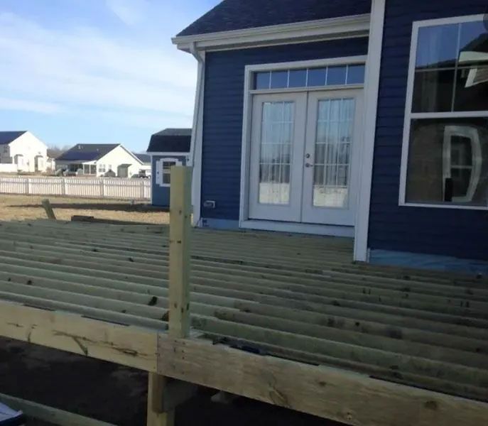 Unfinished wooden deck attached to a blue house with white doors and windows; sunny outdoor setting.