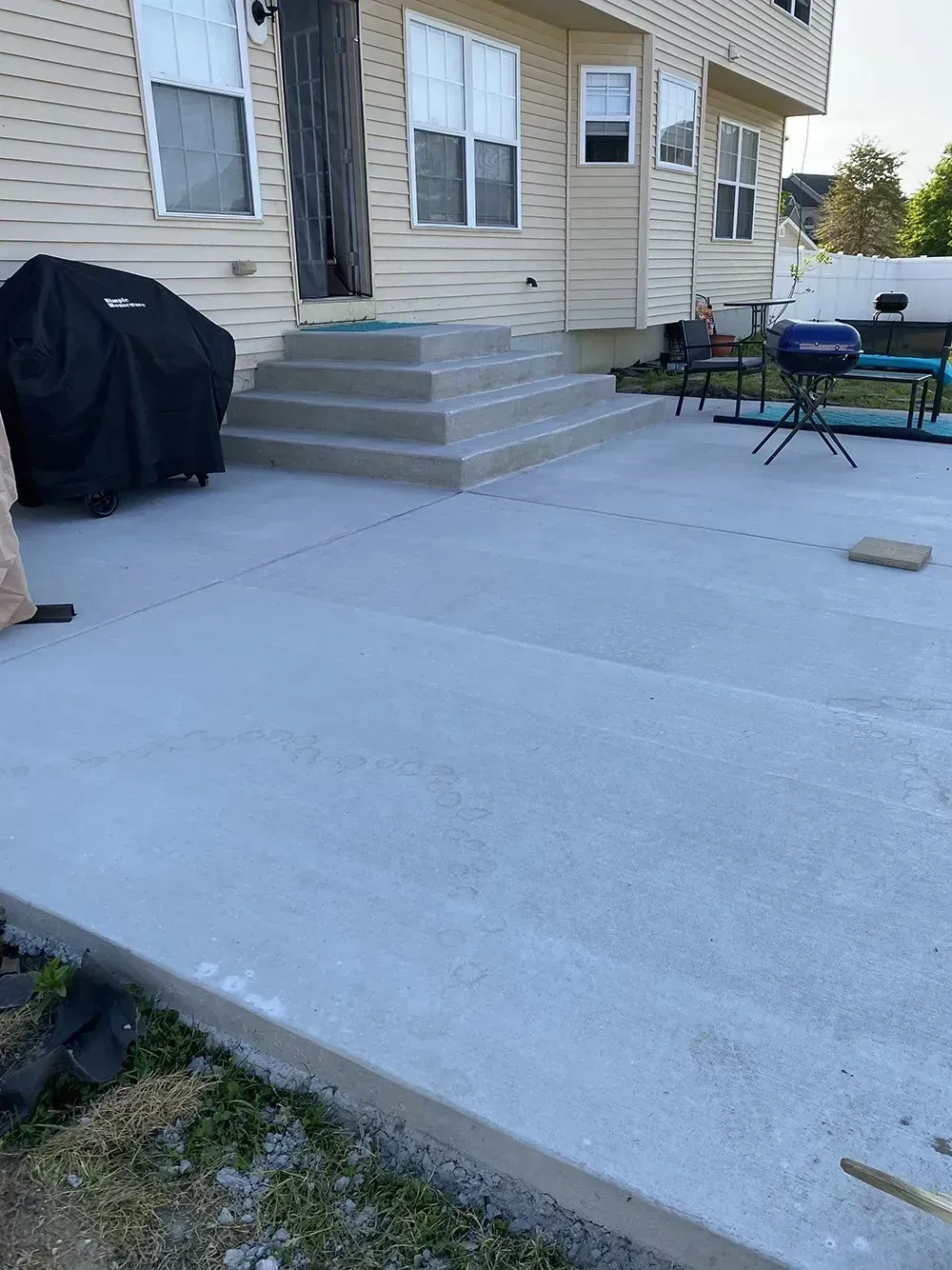 Concrete patio with steps leading to a beige house, covered grill, and outdoor table and chairs.