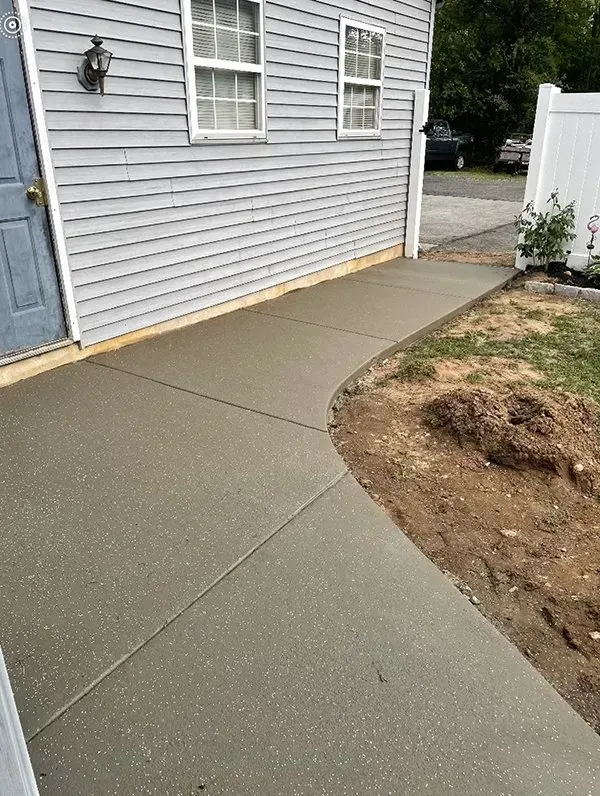 Fresh concrete walkway beside a house, curving around a dirt patch near a white fence.