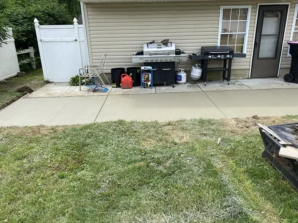 Backyard patio with grill, cooler, and storage items against a beige house, beside a grassy lawn.