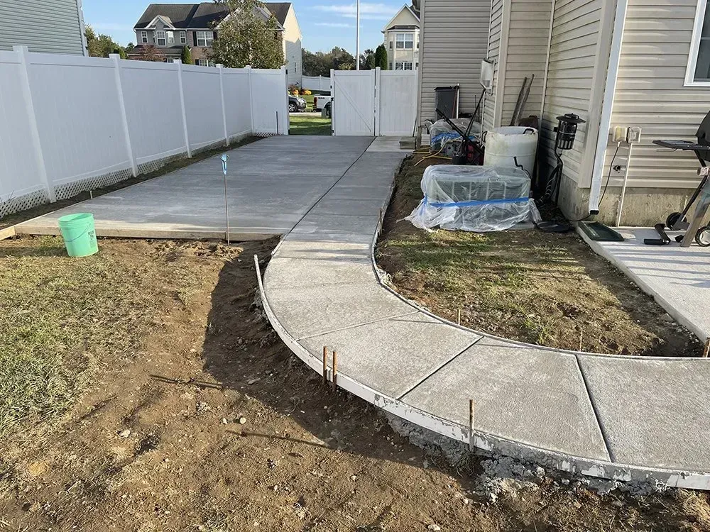Curved concrete walkway beside a house and white fence, with bare dirt landscaping and construction materials.