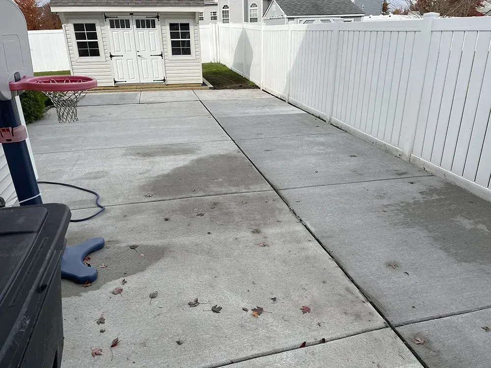 Backyard concrete patio leading to a small white shed, bordered by a white fence and house siding